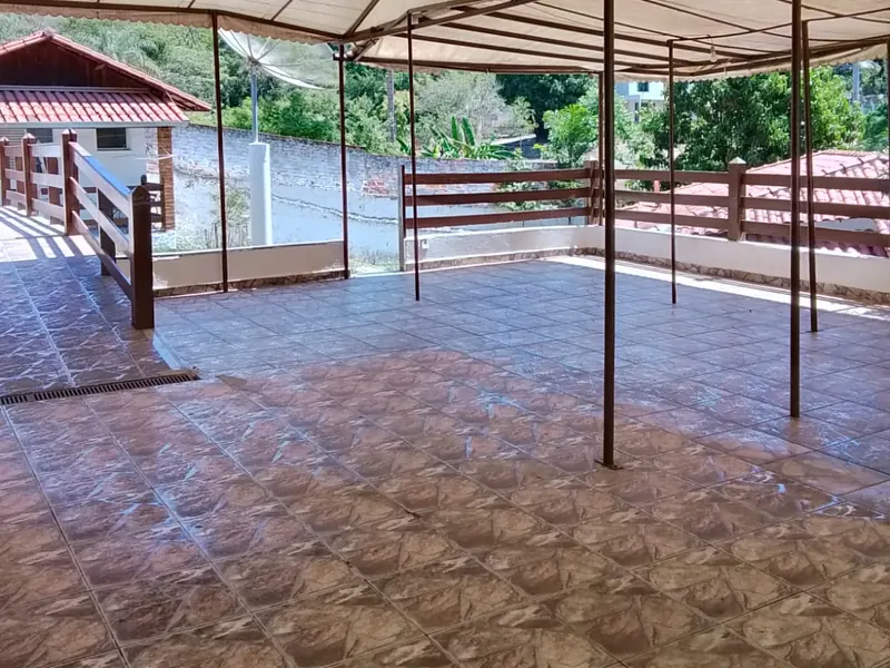 Área ampla coberta por toldo claro, com piso cerâmico, guarda-corpo de madeira e uma antena parabólica ao fundo