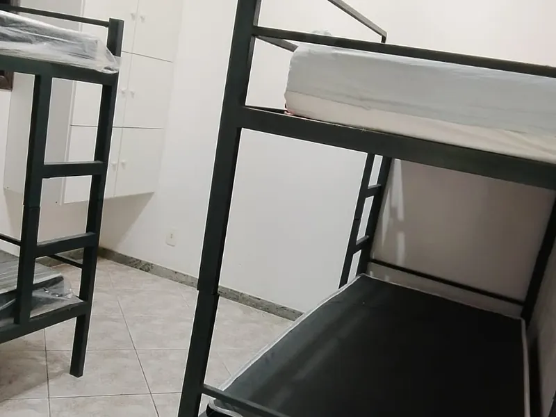 Quarto de paredes brancas com dois beliches de metal escuro e armários brancos ao fundo